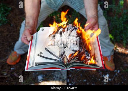 Man holds an open book engulfed in bright orange flames outdoors, depicting a dramatic and symbolic act of book burning, censorship, and the destructi Stock Photo