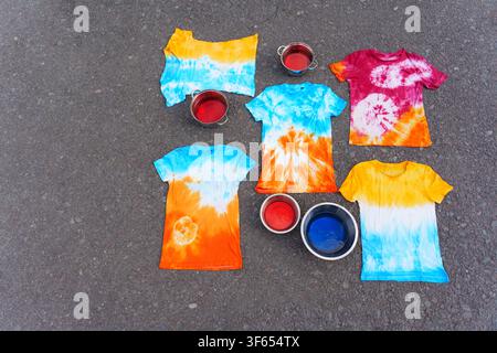 Brightly colored handmade tie-dye t-shirts laid out on asphalt ground next to bowls of liquid dye, showcasing creative DIY craft projects and vibrant Stock Photo
