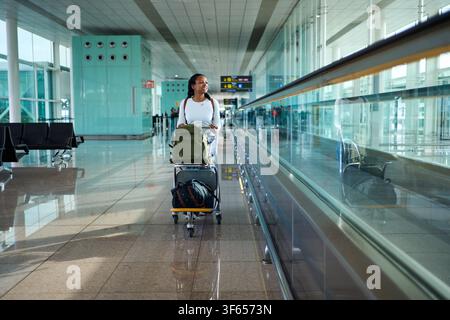 Woman smiling moving through airport terminal with luggage cart. Stock Photo