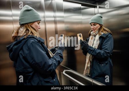 A woman stands in an elevator looking at her smartphone while wearing a coat and hat. It is winter. Stock Photo