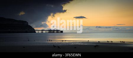 Dramatic sunset over the sea at Llandudno beach, showing the historic Victorian pier silhouetted against the headland of the Great Orme. Panoramic Stock Photo