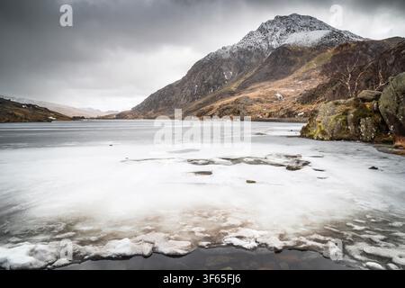 Dramatic winter view looking across the frozen ice of Llyn Ogwen toward the snow-capped peak of Tryfan mountain under an overcast sky. Winter weather Stock Photo