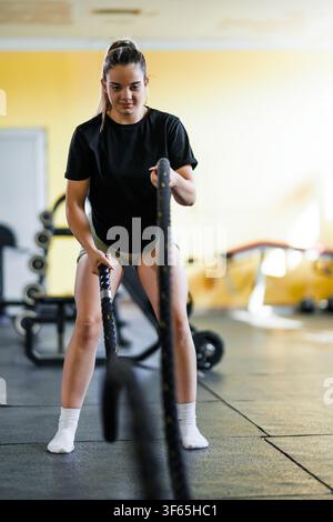 Woman exercises with battle ropes in a gym during daytime showing strength and focus using fitness equipment on black mats Stock Photo