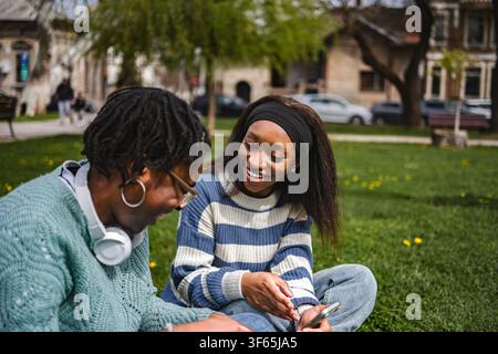 Two young friends sitting on grass using smartphone and sharing joyful moment outdoors Stock Photo