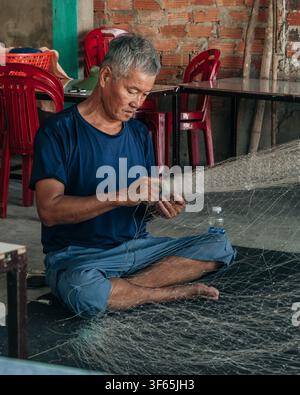 Hoi An, Vietnam, July 15, 2026. A grey-haired man sits on a black mat inside a rustic room with exposed brick walls, carefully working on a large whit Stock Photo