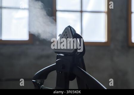 Handheld garment steamer emitting a plume of white steam on a black clothes hanger in a modern industrial setting. Stock Photo
