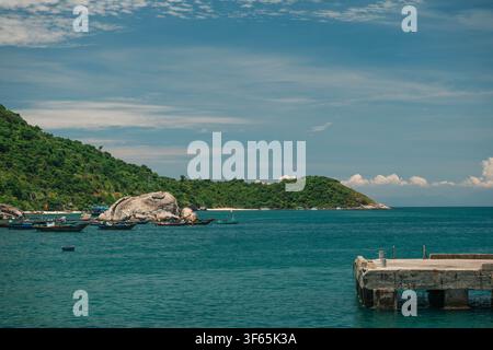 A concrete dock stretches into calm turquoise ocean water near a green hillside shoreline dotted with small anchored vessels under a blue sky. Stock Photo