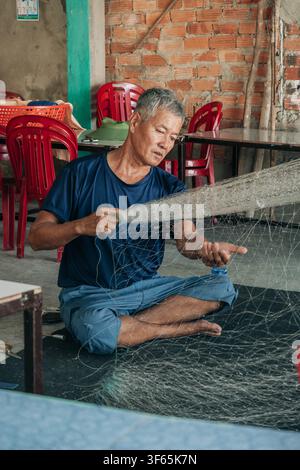 Hoi An, Vietnam, July 15, 2026. A grey-haired man sits on a black mat inside a rustic room with exposed brick walls, carefully working on a large whit Stock Photo