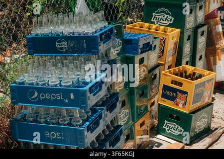 A colorful stack of plastic boxes containing empty glass drink containers rests near a wire fence and styrofoam packages on a bright sunny day. Stock Photo