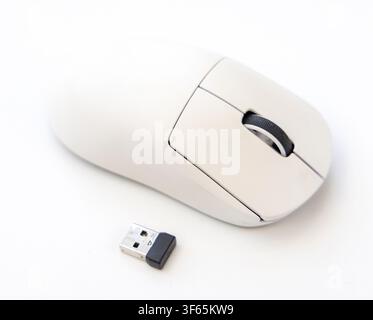 A white wireless computer mouse is placed on a white surface alongside a small black and silver USB receiver Stock Photo