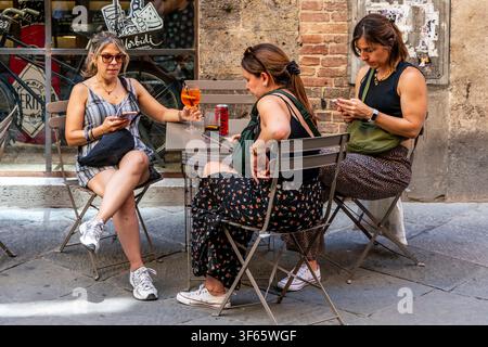 Three Women Sitting Outside A Cafe Drinking Soft Drinks and Looking At Their Phones, Siena, Tuscany, Italy Stock Photo