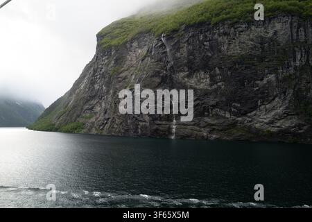 Seagull flying above Geirangerfjord with waterfalls on the cliffs and a ship's wake below, Norway Stock Photo