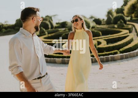 A joyful couple walks hand in hand through an elegant garden. They wear stylish outfits, radiating happiness under the warm sun. Stock Photo