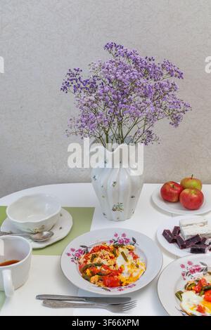 Vertical close up of a white kitchen table served with delicious fried eggs, fresh apples, oatmeal porridge and a flower vase at morning. Stock Photo