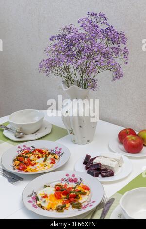 Vertical close up of a white kitchen table served with delicious fried eggs, fresh apples, oatmeal porridge and a flower vase at morning. Stock Photo
