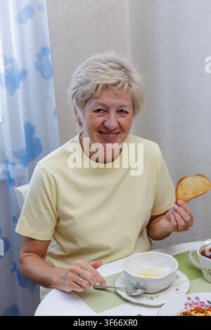 Portrait of a smiling mature woman holding a slice of toasted bread while having healthy porridge and eggs for morning breakfast at home. Stock Photo