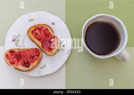 Top angle view of crispy toasts with sweet red raspberry jam and a cup of black coffee served on a green placemat for breakfast. Stock Photo