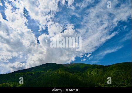A wide landscape photograph capturing a dramatic, dynamic sky filled with large, billowing white and wispy clouds stretching across a vibrant blue background. Below, rolling mountain slopes covered in dense, dark green evergreen forests fill the lower portion of the frame, creating a striking contrast between the earth and sky. Stock Photo