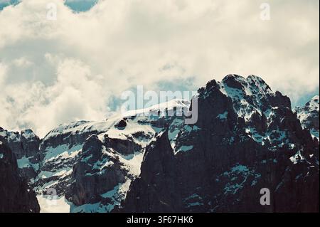A dramatic landscape photograph capturing rugged, dark, rocky mountain peaks partially dusted with snow, rising sharply against a soft, overcast sky filled with dense white and grey clouds. The deep contrast emphasizes the sharp textures and jagged contours of the high alpine terrain. Stock Photo