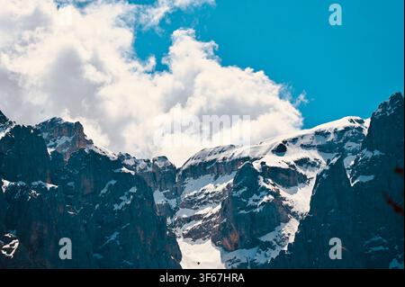 A dramatic landscape photograph capturing rugged, dark, rocky mountain peaks partially dusted with snow, rising sharply against a soft, overcast sky filled with dense white and grey clouds. The deep contrast emphasizes the sharp textures and jagged contours of the high alpine terrain. Stock Photo