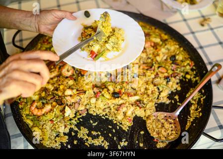 Person scooping delicious mixed rice with shrimp and vegetables from a large black pan on a checkered tablecloth Stock Photo