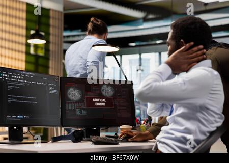Teamworking technicians resolving critical error alert on screen in tech startup office, troubleshooting system warnings. IT specialist and coworker struggling with office equipment failure Stock Photo