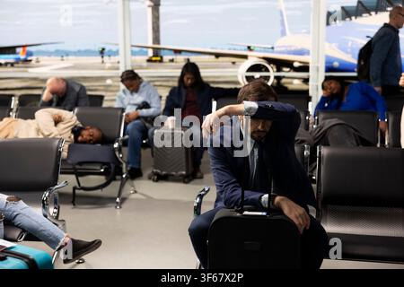 Tired CEO man in airport terminal lounge trying to rest during delayed flight business trip. Exhausted businessman sleeping on trolley handle, waiting for canceled flights to resume Stock Photo