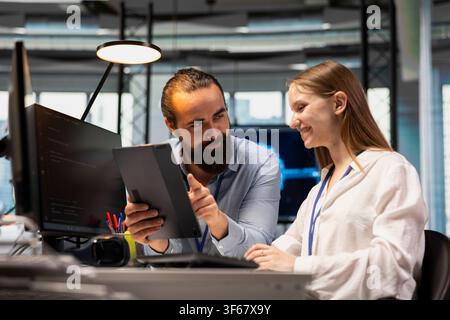 Cheerful technician mentoring young employee in startup office, reviewing code scripts on tablet. Jolly tech company team leader guiding intern doing software development Stock Photo