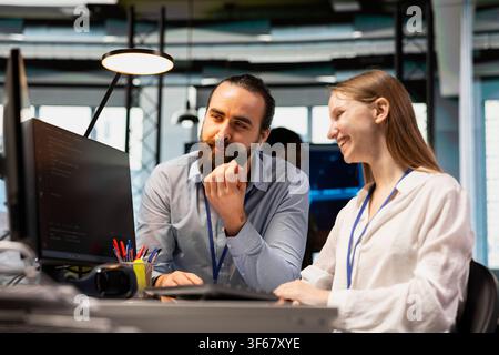 Jolly system administrator mentoring junior dev at desk in office, teaching best coding practices. Smiling tech startup IT specialist guiding female developer intern through project tasks Stock Photo