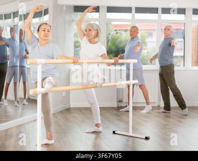 Elderly women with group of dancers practicing ballet pose grand batman near ballet barre in dancing hall Stock Photo