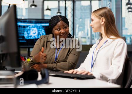 Cheerful IT specialist teaching intern to use artificial intelligence. African american woman guiding junior dev through AI tool integration, mentoring her on using machine learning APIs Stock Photo