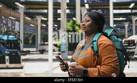 Close up of woman using smartphone in bus station while enjoying coffee. Solo passenger carries takeaway drink cup and mobile phone during coach intercity departure Stock Photo