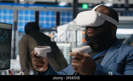 Black photovoltaics factory expert uses VR glasses to assembly digital solar cells, working with robotic arms for solar panels placement. Engineer uses machinery with virtual reality. Camera B. Stock Photo