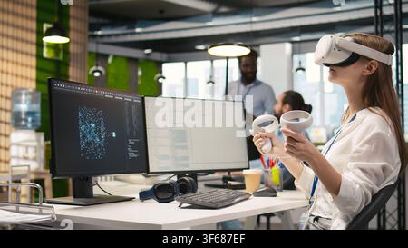Female technician wearing VR goggles coding with AI language model assistance in startup office. Young woman using virtual reality integrating LLM assistant suggestions into code editor, camera A Stock Photo