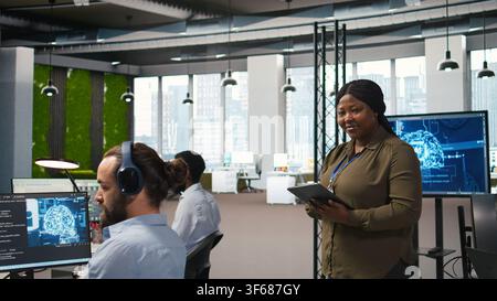 Smiling female engineer next to office coworkers developing software applications, integrating AI automation. Happy woman next to developers at work debugging applications, camera A Stock Photo