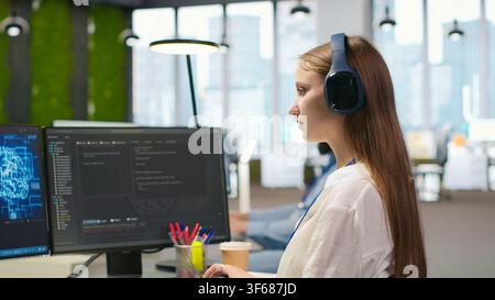 Junior dev developing software applications, optimizing code for performance, listening music. Young female engineer wearing headphones in office using AI to resolve bottlenecks, camera A Stock Photo