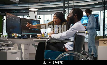 Startup office engineer in wheelchair talking with colleague, developing artificial intelligence models. IT specialist with paraplegia and coworker running simulations for AI forecasting, camera A Stock Photo