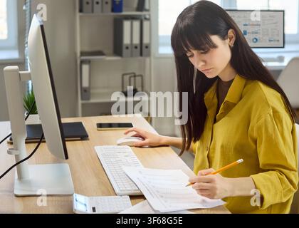 Focused woman accountant reviewing budget paperwork at desktop computer in office Stock Photo
