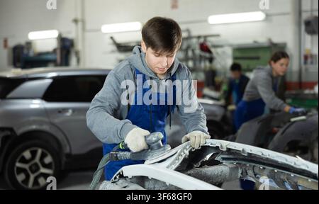 Male mechanic prepares car for painting in workshop - using grinding machine he sands bumper before painting Stock Photo