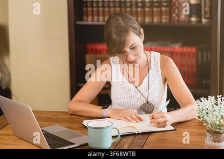 Woman with glasses writing in a notebook at a wooden home workspace with laptop and coffee mug, representing remote work, planning and daily productiv Stock Photo