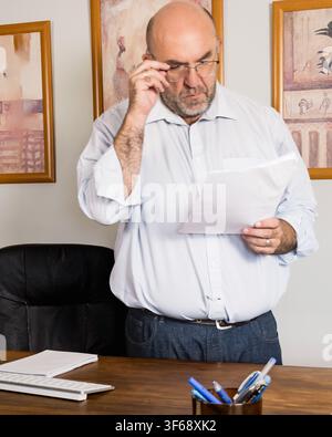Mature businessman stands beside his desk, adjusts his glasses and examines printed documents in a professional office, conveying careful review and d Stock Photo