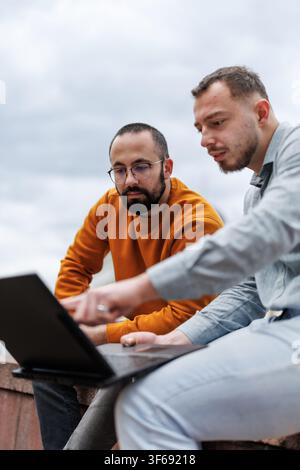 Two colleagues collaborating outdoors on a laptop discussing a project, sharing ideas and focused teamwork in a casual business setting Stock Photo