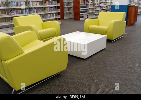 A wide interior shot of a bright, modern public library or study center. The space features comfortable yellow armchairs arranged around a coffee tabl Stock Photo