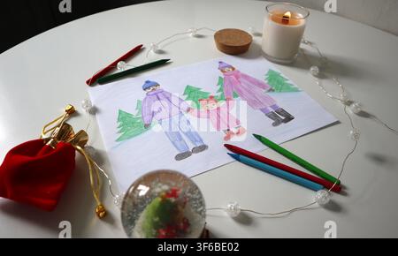 A family is drawn on a paper holding hands in winter clothing. Colored pencils are present on the table along with decorations and a lit candle. The s Stock Photo