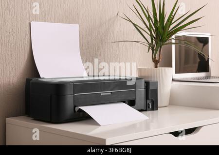 Black inkjet printer with blank paper on a white desk in a home office setting with a laptop and plant, a laptop and a potted plant are visible in the Stock Photo