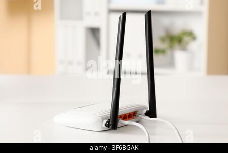 White wireless router with two antennas and connected ethernet cables on a clean white desk in an office setting, the background shows a blurred offic Stock Photo