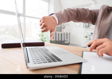 Businessman giving a thumbs down gesture while working on a laptop at a modern office desk, expressing disapproval or dissatisfaction Stock Photo