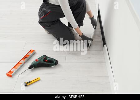 Worker installing new baseboard trim along a white wall on light wood laminate flooring with tools nearby Stock Photo