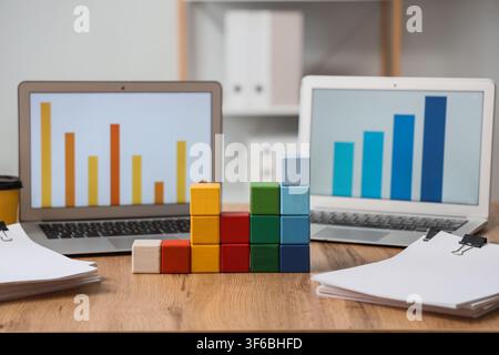 Colorful wooden blocks forming a bar graph in front of laptops displaying data charts on a wooden desk Stock Photo