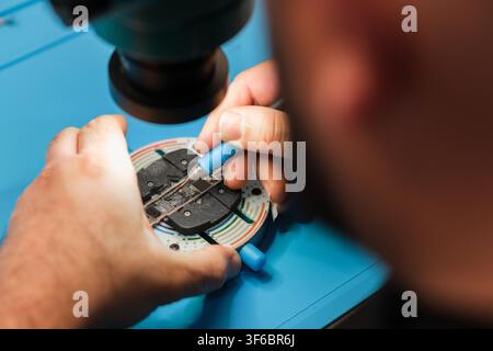 Technician repairs electronic circuit board under microscope with precision tools on a blue workbench in a laboratory setting Stock Photo